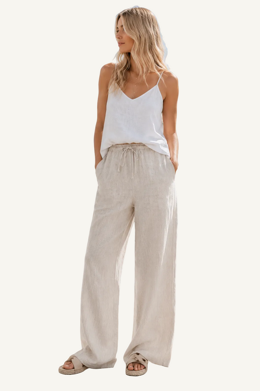 Woman in white top and beige pants standing on a sidewalk with palm trees and buildings in the background.