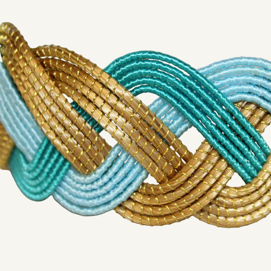 A close-up of a braided woven bracelet featuring interwoven strands of metallic gold, turquoise, and light blue.