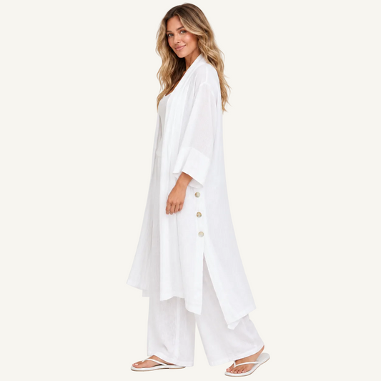 A woman modeling a white long-sleeve button-front tunic with wide legs, paired with matching white pants and white sandals, against a plain off-white studio background.