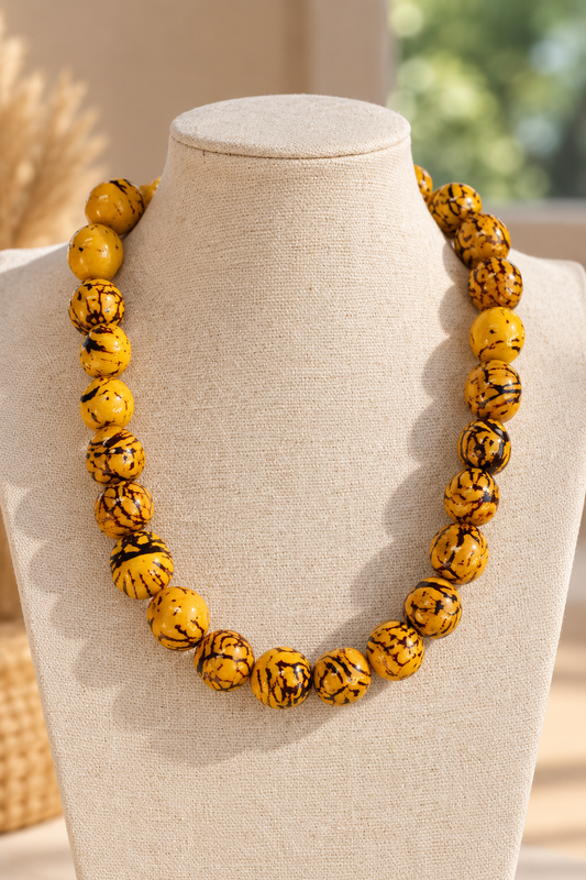 A chunky beaded necklace made of round yellow beads with black marbled swirls, displayed on a beige fabric jewelry bust against a softly blurred warm indoor background.