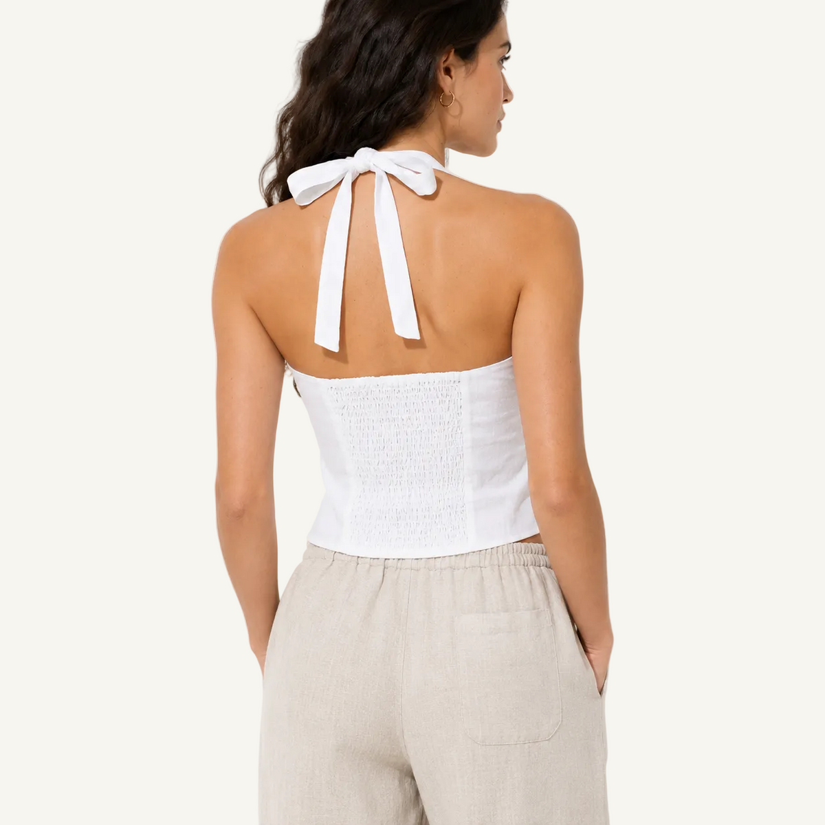 A woman shown from the back wearing a white halter-style smocked crop top with long tie straps at the neck, paired with light beige pants, against a plain off-white background.
