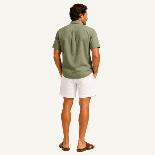 A man shown from the back wearing an olive green short-sleeve button-up shirt and white linen shorts, paired with brown leather slide sandals, against a solid off-white background.