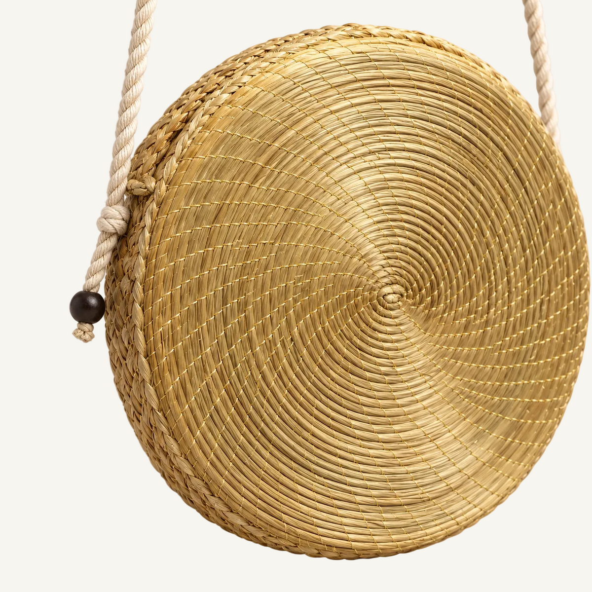 A round woven straw bag with a spiral pattern, featuring a cream-colored rope shoulder strap with a dark wooden bead accent, set against a plain light gray background.