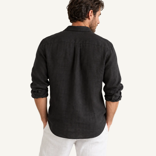 A man viewed from the back wearing a dark charcoal linen button-down shirt with rolled-up sleeves and white trousers, set against a plain off-white studio background.
