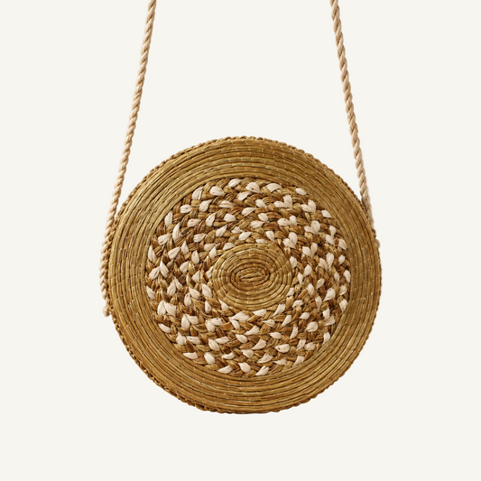 Round woven straw shoulder bag with a rope strap, featuring a spiral pattern of natural tan and off-white raffia, set against a warm beige background with soft palm leaf shadows.