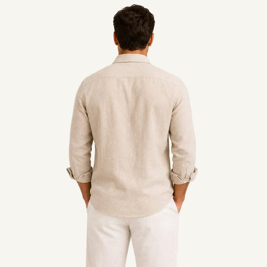 A man viewed from behind wearing a light beige linen button-down shirt with rolled-up sleeves and white trousers, standing against a neutral light beige background.
