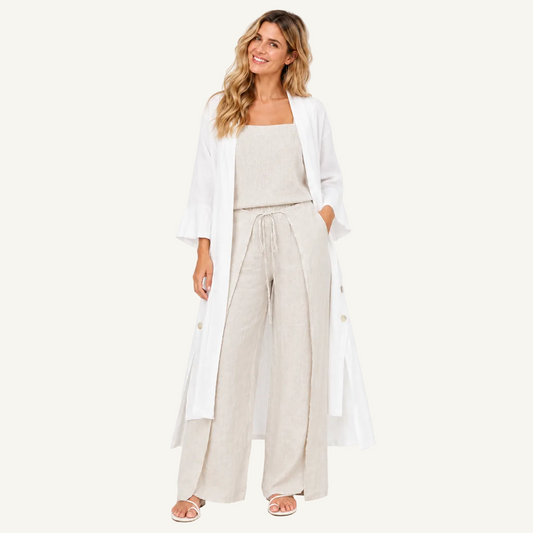 A woman modeling a light beige linen set consisting of a sleeveless top and wide-leg drawstring pants, styled with a long white open-front cardigan and white sandals against a plain white studio background.