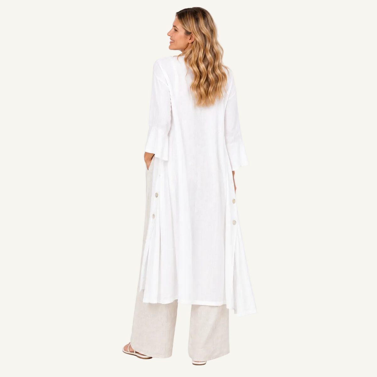 A woman is shown from the back wearing a long white tunic with side buttons, layered over wide-leg beige trousers, against a plain white studio background.