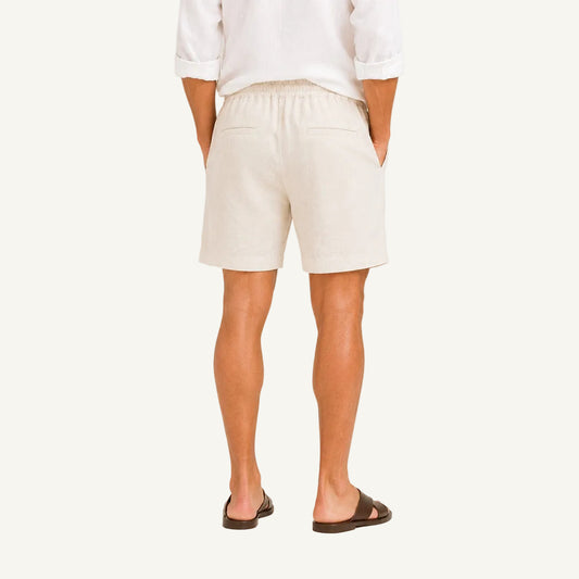 A man viewed from behind wearing off-white linen shorts, a white long-sleeve shirt, and dark brown slide sandals, set against a plain light beige studio background.