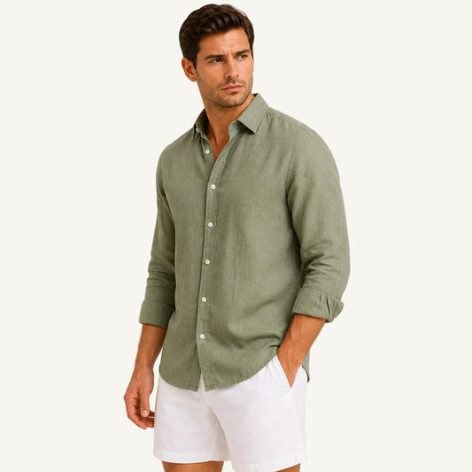 A man wearing a sage green linen button-down shirt and white shorts, standing against a plain light beige studio background.