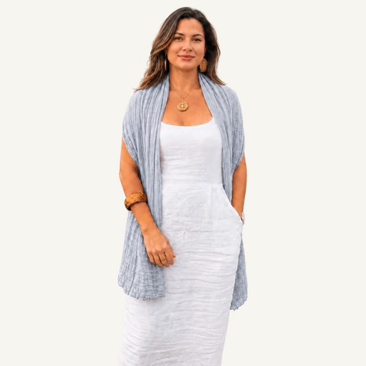 A woman modeling a light blue linen shawl draped over a white sleeveless dress, standing against a neutral beige textured background.
