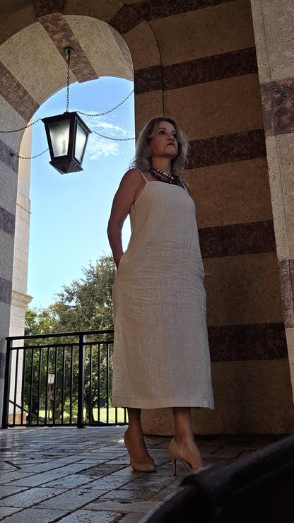 A woman wearing a sleeveless, knee-length cream linen dress and beige heels stands under a stone archway with a hanging lantern, looking upward.