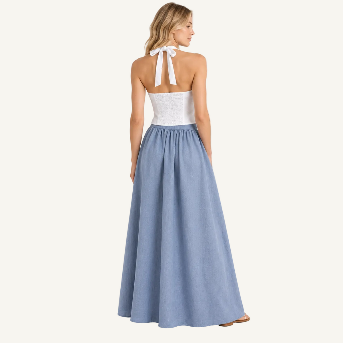 A woman stands with her back to the camera wearing a sleeveless maxi dress with a white lace halter-style bodice and a flowing light blue skirt, photographed against a plain light gray studio background.