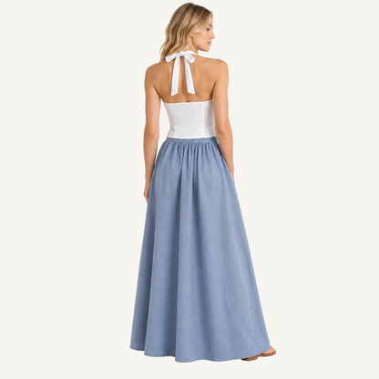 A woman stands with her back to the camera wearing a sleeveless maxi dress with a white lace halter-style bodice and a flowing light blue skirt, photographed against a plain light gray studio background.