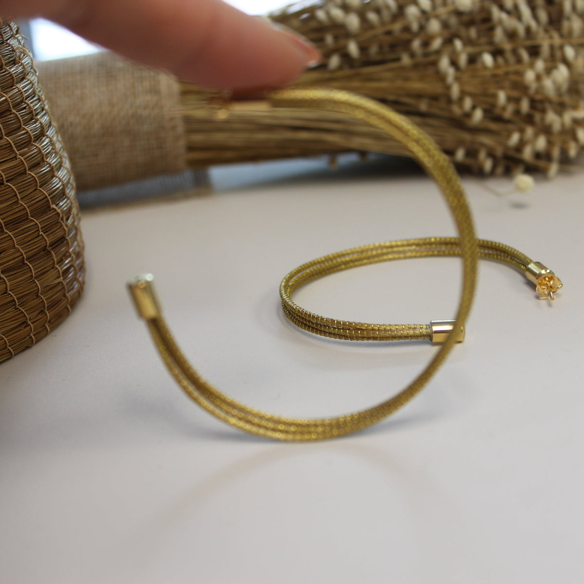 A gold-colored braided elastic bracelet with a small star charm, shown on a white surface with a hand pointing at it.