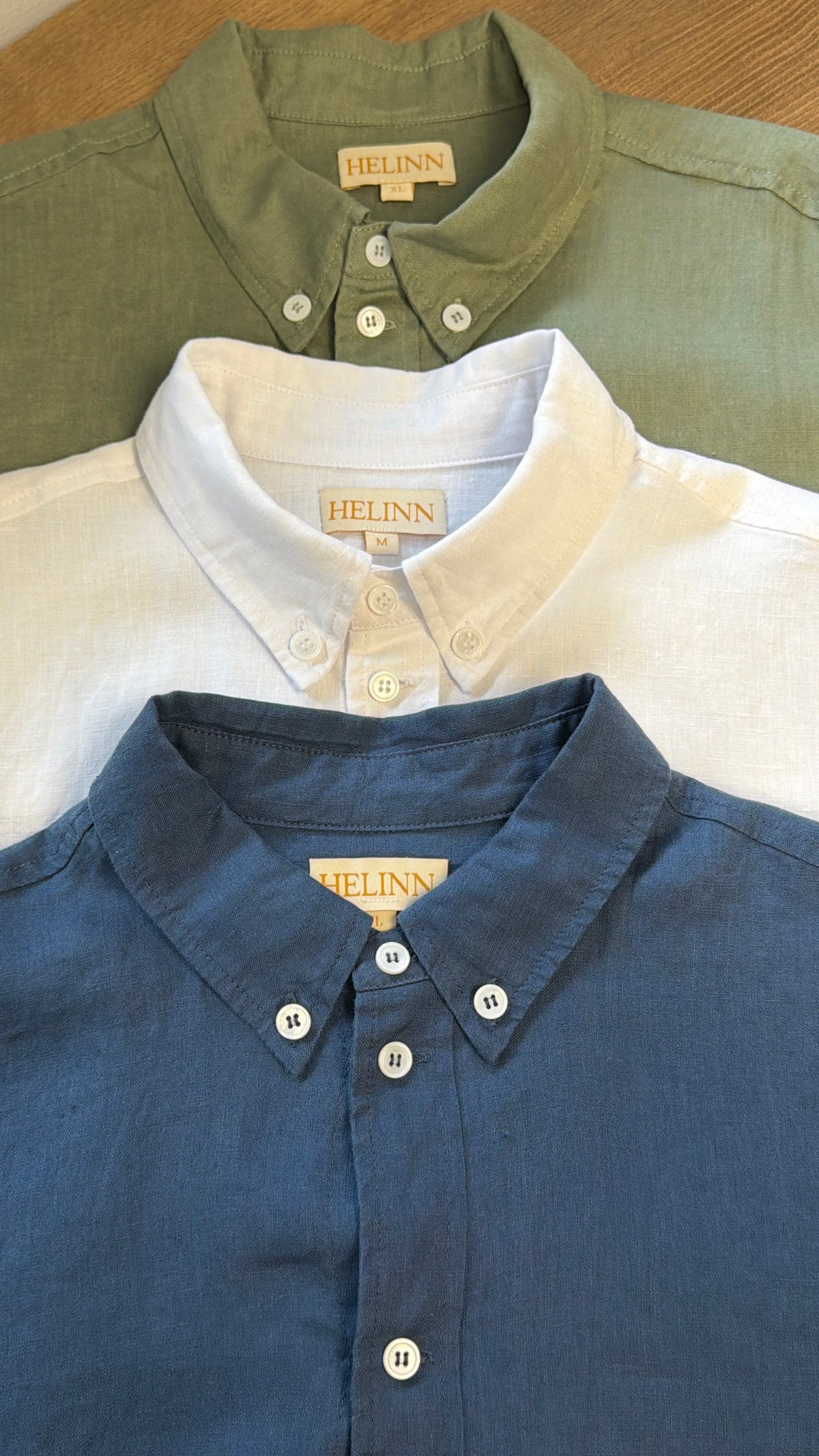 Three Helinn button-down linen shirts in sage green, white, and slate blue, arranged in a stacked flat-lay on a wooden surface.