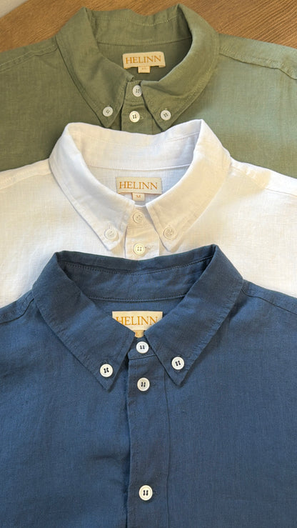 Three Helinn button-down linen shirts in sage green, white, and slate blue, arranged in a stacked flat-lay on a wooden surface.