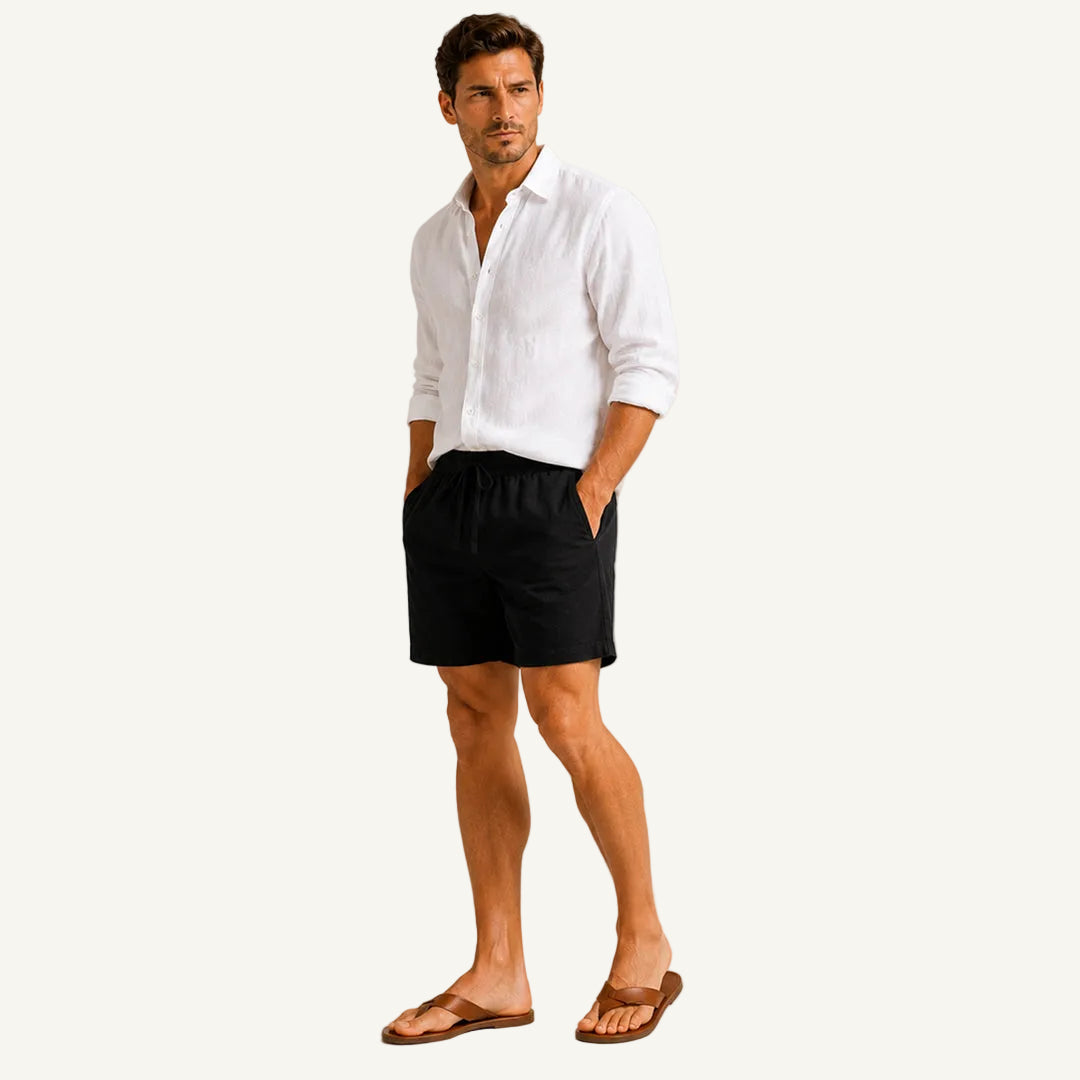 A man modeling a white long-sleeve linen shirt, black drawstring shorts, and brown leather sandals against a neutral beige studio background.