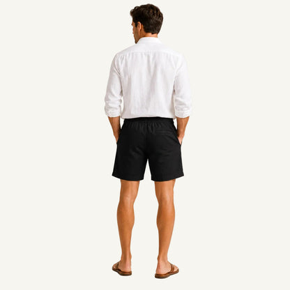 A man viewed from behind wearing a white long-sleeve button-up shirt, black shorts, and brown sandals, standing against a plain light beige studio background.