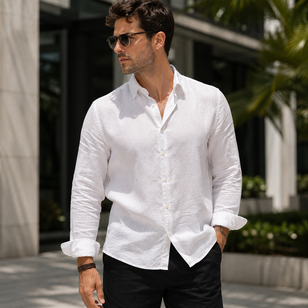 A man wearing a white long-sleeve linen button-down shirt and black pants, standing outdoors in front of a modern building with greenery.