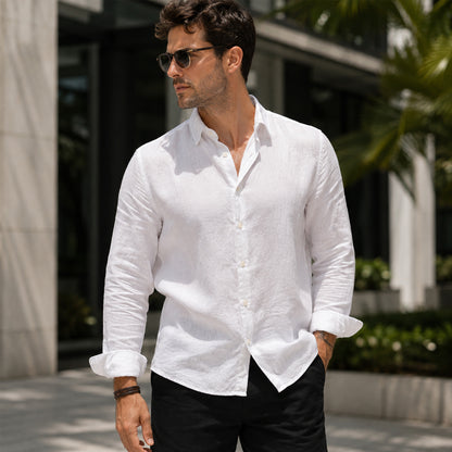 A man wearing a white long-sleeve linen button-down shirt and black pants, standing outdoors in front of a modern building with greenery.