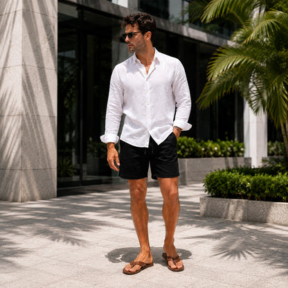 A man wearing a white linen long-sleeve button-down shirt, black shorts, and brown sandals stands outdoors in front of a modern building with palm trees.