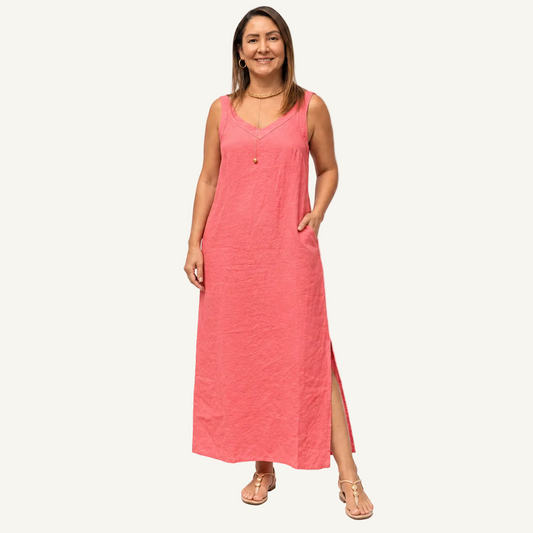 A woman modeling a sleeveless coral pink maxi dress with a V-neckline, side pockets, and a front slit, photographed against a clean white studio background.