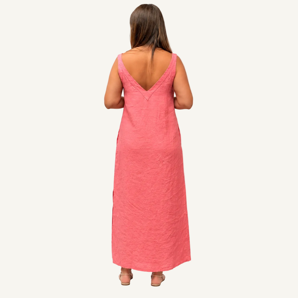 A woman viewed from behind wearing a linen sleeveless coral pink maxi dress with a deep V-neckline and side slits, standing against a plain white studio background.