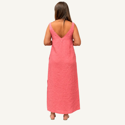A woman viewed from behind wearing a linen sleeveless coral pink maxi dress with a deep V-neckline and side slits, standing against a plain white studio background.