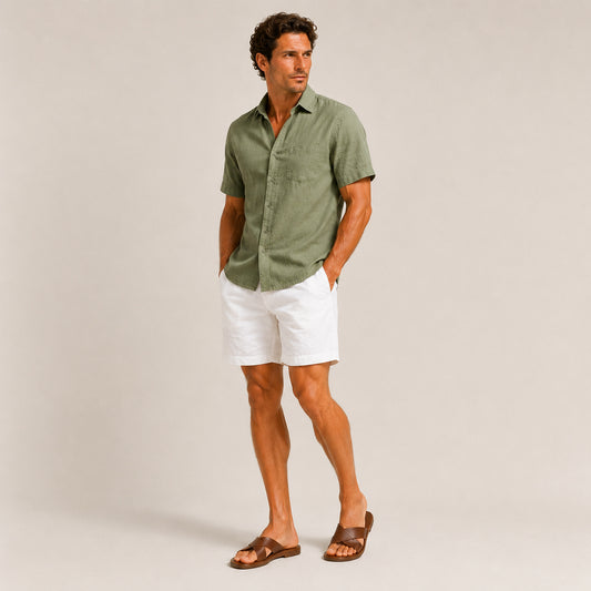 A man modeling a short-sleeve olive green linen button-down shirt, white linen shorts, and brown leather slide sandals against a light beige studio background.