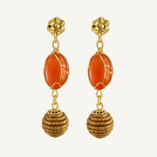 A pair of gold-plated drop earrings featuring a polished orange carnelian stone in the center and a textured gold beaded sphere dangle below, set against a plain white background.