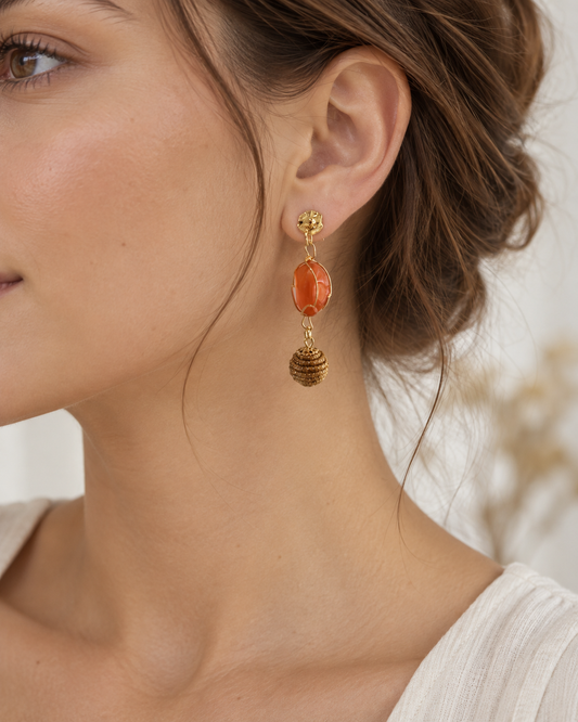 A close-up of a woman's ear wearing a gold dangle earring with a polished orange stone and a textured gold bead drop.