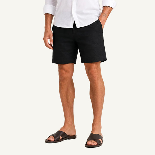 A man wearing black linen shorts, a white button-down shirt, and dark brown leather slide sandals, standing against a light gray studio background.