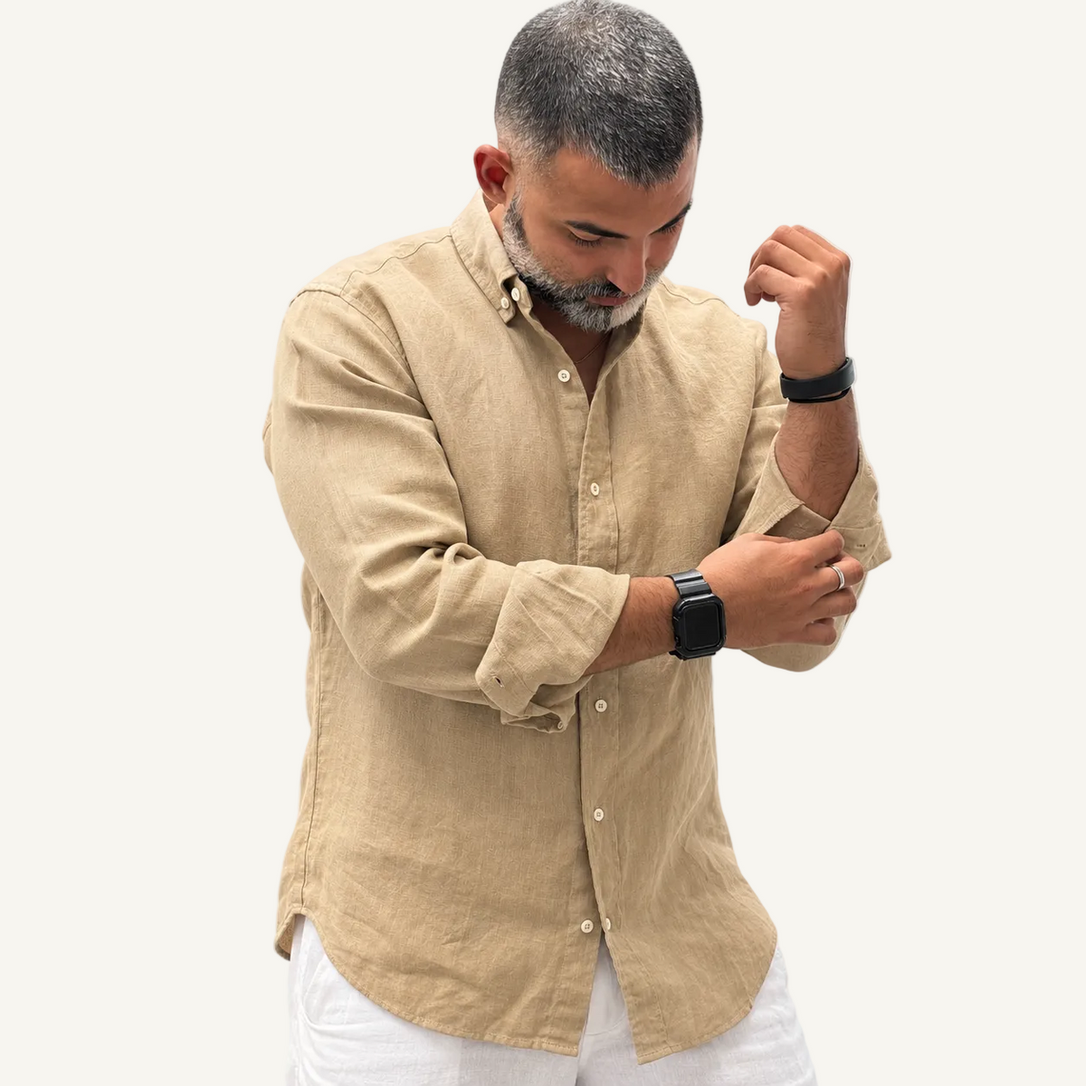 A man wearing a light beige linen button-down shirt and white pants, adjusting his rolled-up sleeve while looking down. He is wearing a black smartwatch on his wrist.