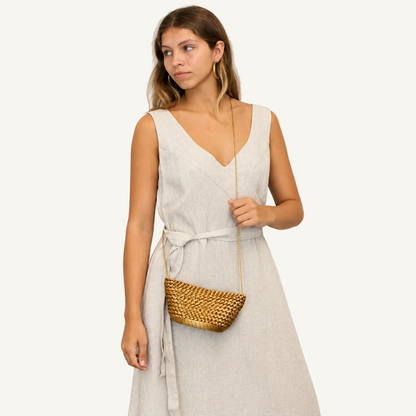 A woman wearing a light beige linen wrap dress with a tie waist, holding a small woven gold crossbody bag, set against a plain light gray background.