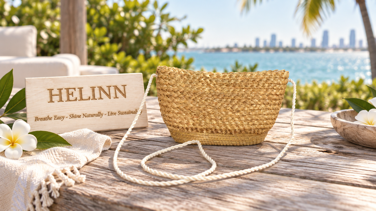 A woven straw shoulder bag with a cream braided strap sits on a wooden table outdoors, with a tropical ocean and city skyline in the background. A wooden sign reading HELINN is placed beside it, along with white flowers and a folded cream textile.