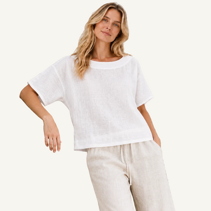 A woman wearing a loose white short-sleeve linen top and light beige drawstring pants, leaning against a textured light stone wall.