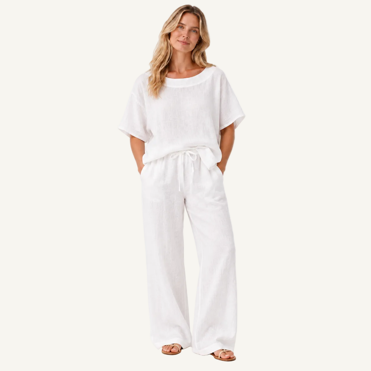 A woman modeling a white linen two-piece set consisting of a short-sleeve tunic top and matching wide-leg pants, styled with brown leather sandals against a plain white studio background.