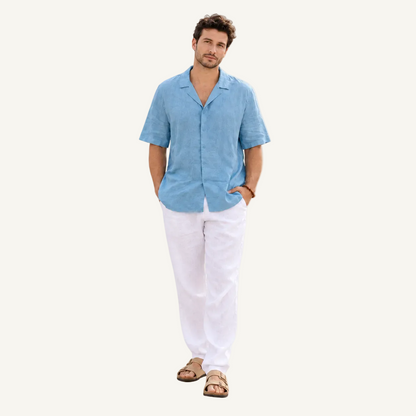 A man modeling a light blue short-sleeve linen button-up shirt and white linen pants, standing against a solid off-white background.