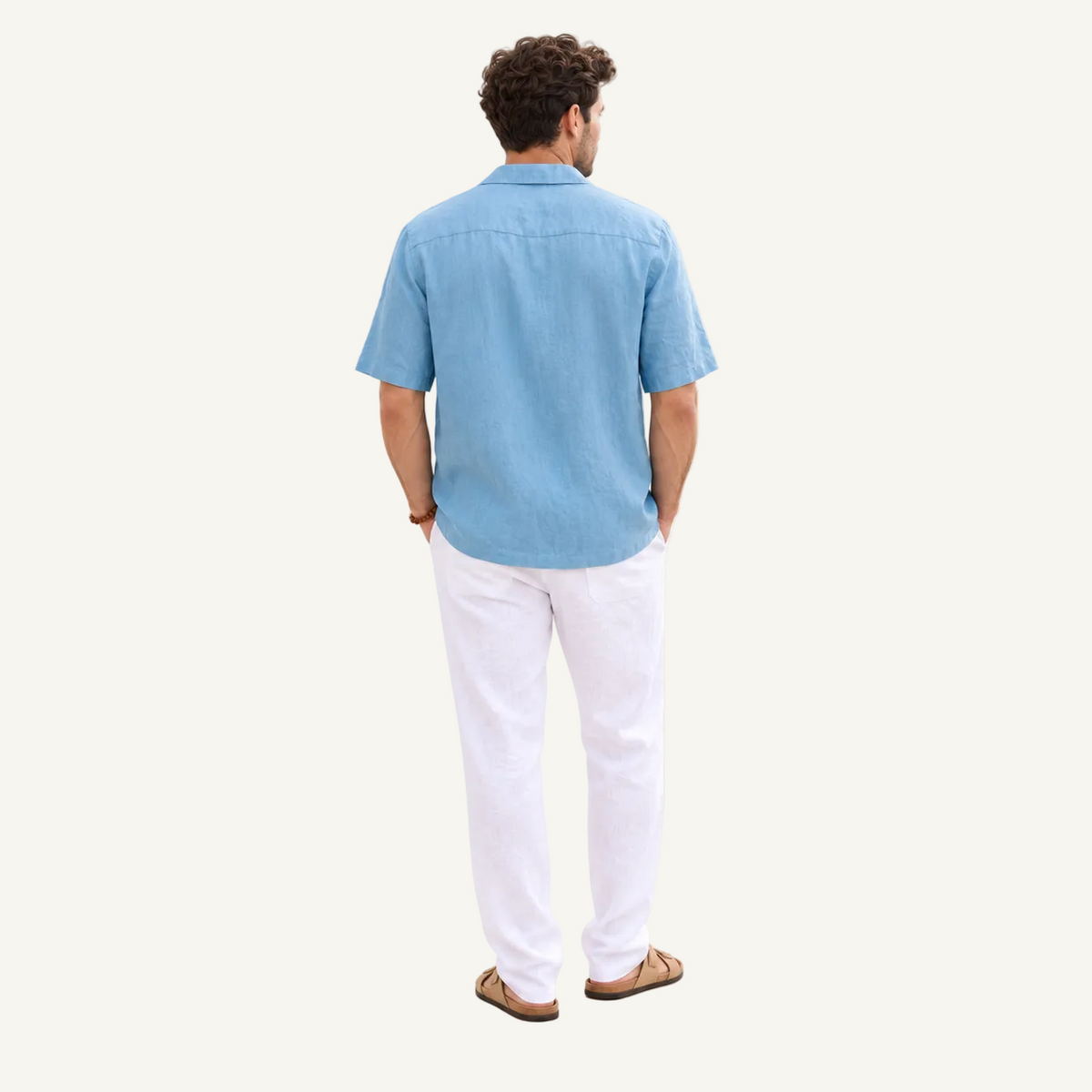 A man viewed from behind wearing a light blue short-sleeve button-up shirt, white trousers, and brown sandals, standing against a plain off-white studio background.