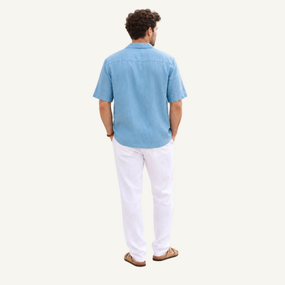 A man viewed from behind wearing a light blue short-sleeve button-up shirt, white trousers, and brown sandals, standing against a plain off-white studio background.